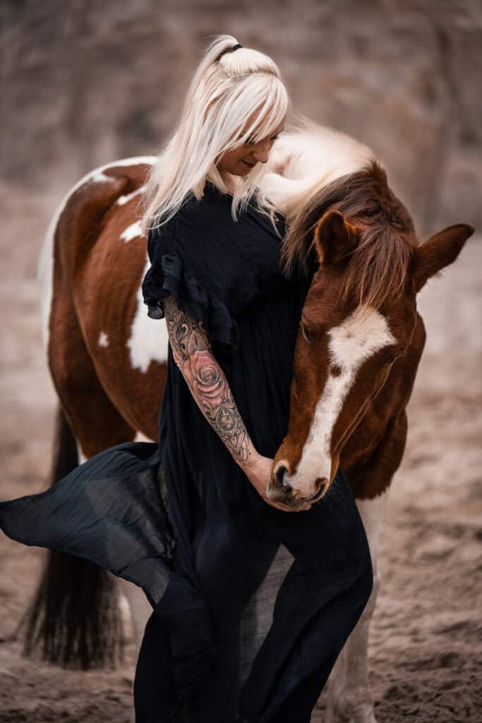 Frau mit langen blonden Haaren und Tattoo umarmt braunen Pferdekopf.
