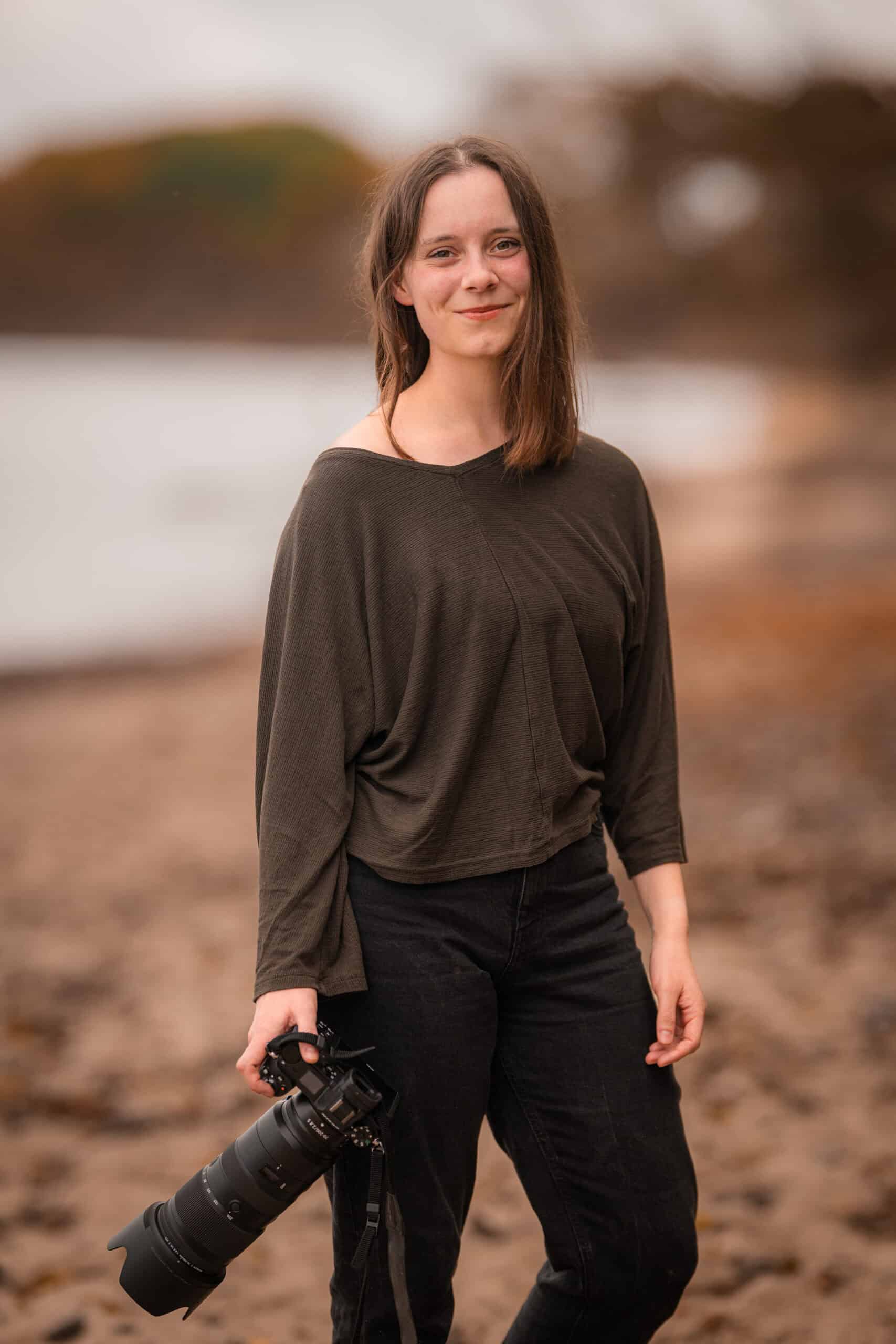 Alena Süßenbach, professionelle Fotografin, mit Kamera in der Hand, steht am Strand bei Sonnenunterg.