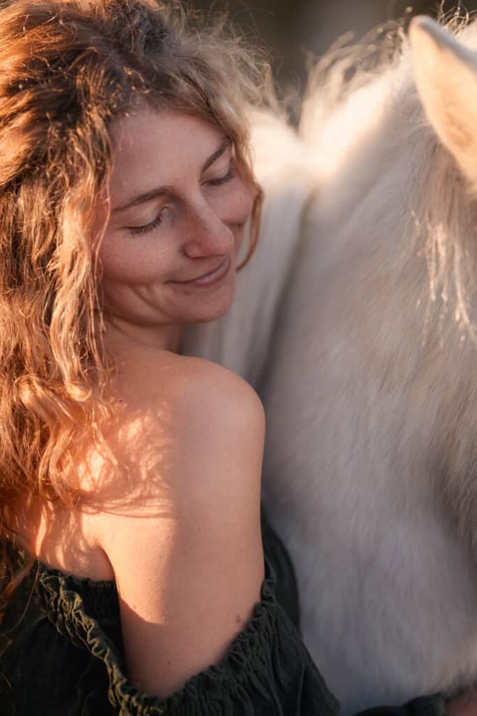 Frau mit lockigem Haar umarmt weißes Pferd bei Sonnenuntergang.